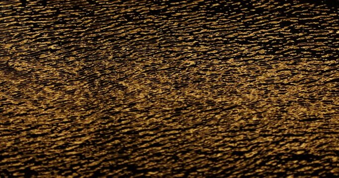 Cinematic close-up of dark rippling water covered with shimmering golden reflections, creating an abstract, luxurious and atmospheric texture with dynamic wave patterns and high-contrast highlights