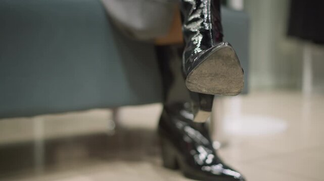Close up of woman foot in black shiny boots with high heels showing slight movement and texture detail. background is softly blurred suggesting indoor setting like store or salon. Lighting