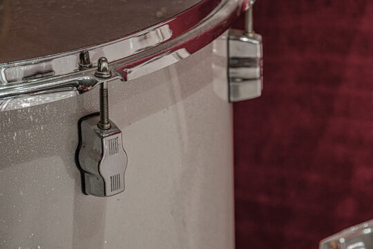 Close-up view of drum set hardware, focusing on metallic polished tension rods and lugs, with a white worn drum head against deep red textured fabric background, bright focused lighting, high contrast