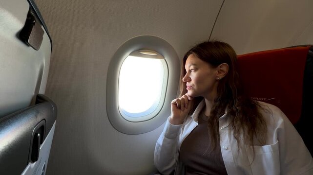 Woman looks pensively out of airplane window, seated comfortably in a modern aircraft cabin, contemplating the view outside