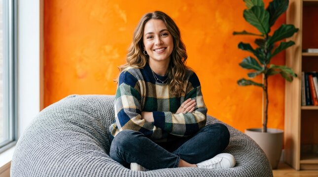 Portrait of a smiling young woman sitting cross-legged in a grey beanbag chair against a vibrant orange wall in a modern apartment.