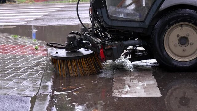 Close-up of a mechanical street sweeper cleaning a wet asphalt road during rainy weather. Rotating brush removes dirt, leaves and debris from the road surface, showing municipal cleaning, urban mainte