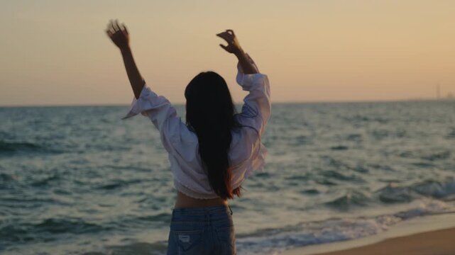 Young woman on beach waves hand in goodbye gesture at sunset, enjoying peaceful evening view of ocean waves, wearing white shirt and casual denim jeans during summer vacation travel.