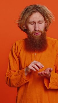 Need some more, please give me. Caucasian man showing a little bit gesture with sceptic smile showing minimum sign, measuring small size begging help. Redhead guy isolated on orange studio background