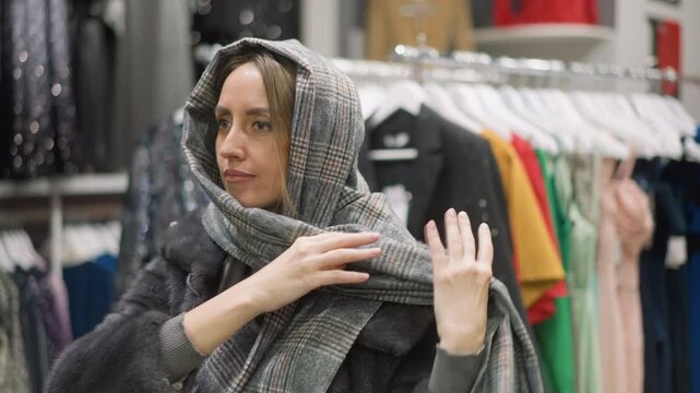 Close up of woman trying on winter clothing in store adjusting and draping gray plaid scarf over her head while wearing dark fur coat. Clothing racks with various garments hang behind her
