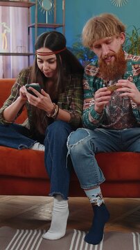 Family married hippie couple man woman using credit bank card and smartphone while transferring money, purchases online shopping, order food delivery at home. Husband and wife together on sofa in room