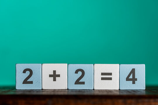 Simple math equation two plus two equals four on wooden toy blocks. Row of light blue and white wooden blocks spelling out basic arithmetic addition formula 2 plus 2 equals 4 on green background