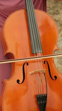 Talented woman's hand moving a bow across the strings of a shiny wooden cello, creating beautiful music during an outdoor concert rehearsal on a bright, sunny afternoon. Vertical video