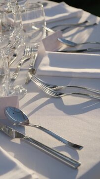 Beautifully decorated table set for an outdoor wedding banquet, with elegant cutlery, glasses, and white napkins creating shadows on the tablecloth in the warm sunlight of a perfect day. Vertical vide