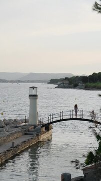 Scenic view of a young woman in a swimsuit walking across a small arched footbridge by the sea, with a quaint lighthouse and a calm coastline visible during a serene, gentle sunset. Vertical video