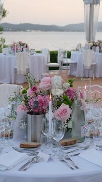 Beautifully decorated table for a wedding reception or romantic dinner at an outdoor restaurant by the sea, featuring a floral centerpiece with pink roses and a flickering candle flame. Vertical video