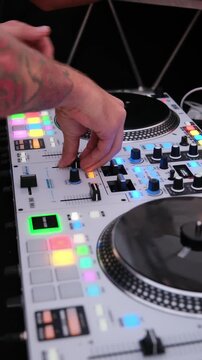 Professional disc jockey with tattoos playing and mixing electronic music on a modern audio controller with turntables, colorful lights, knobs, and faders at a party in a nightclub. Vertical video