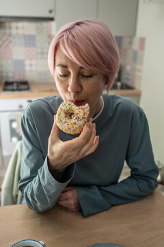 Mature woman eats colorful donut after giving in to intense PMS cravings, showing dietary relapse, sugar addiction and return to sweet pastries despite determined effort to reduce sugar consumption.