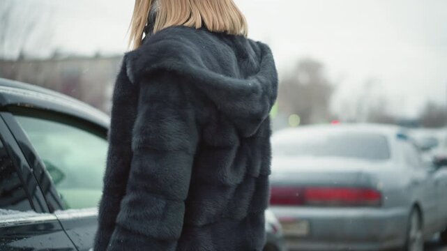 Close up of woman wearing dark fur coat and plaid scarf standing near parked cars in outdoor parking lot. subject remains mostly stationary with slight hand movements surrounded by other