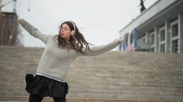 Young woman dances energetically outdoors on stone steps wearing gray knit sweater and black skirt with windblown hair. background features overcast sky modern building facade construction