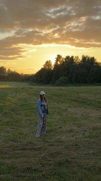 Young woman strolls across a grassy field during sunset, dressed in a denim jacket and patterned pants, with trees and clouds visible