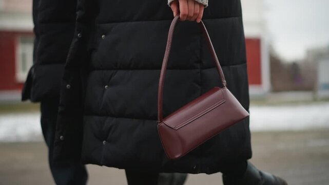 Close up of woman wearing black puffer coat walking outdoors holding maroon handbag. subject moves steadily through urban environment with snow patches on ground and red buildings