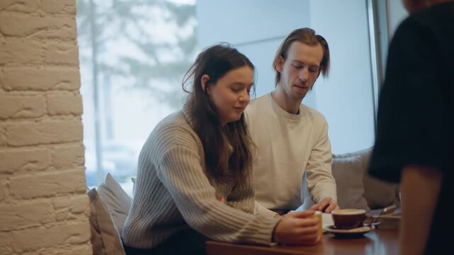 Close up of two people sitting at table in soft lighting interacting with each other and objects on wooden surface. third person briefly appears to serve them beverages or items. setting