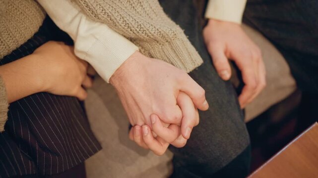 Close up of two people holding hands showing moment of connection and support. One person wears dark pinstriped fabric while other has textured knitwear both are seated on soft indoor