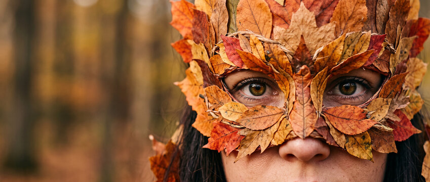 Woman face closeup in mask of autumn leaves in forest. Beautiful mysterious girl in fall foliage crown. Nature fairy and dryad concept. Autumn season and Halloween celebration