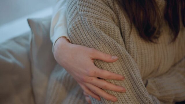 Close up of woman in soft lighting gently holding her arm with visible skin texture and freckles. Woman wears beige knitted sweater against indoor background with neutral tones. focus