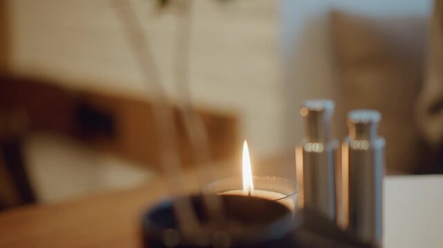 Close up of lit candle on wooden surface with soft warm light and blurred background. Candle flame flickers gently while metal objects stand nearby creating calm indoor atmosphere