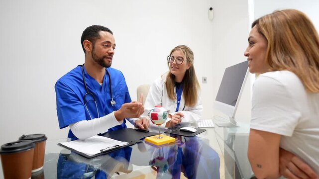Professional male ophthalmologist and a female assistant talking with a patient in a modern clinic office, using an anatomical eyeball model to explain a medical condition or procedure