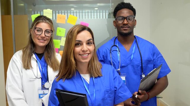 Confident multicultural team of healthcare professionals, including a doctor and two nurses, smiling at the camera while standing in a modern clinic corridor, ready for patient care
