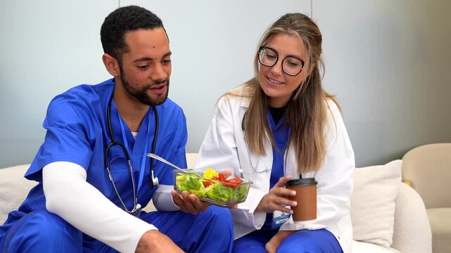 Male and female doctors in uniform sitting on a sofa during their lunch break at the hospital, chatting and enjoying a healthy meal with fresh salad and coffee in the staff room