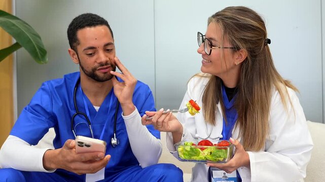 Two diverse colleagues, a male nurse and a female doctor, happily chatting and sharing a healthy salad lunch during a break, while the man also uses his smartphone in the hospital staff room