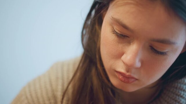 Close up of woman with long brown hair wearing beige knitted sweater softly lit against light blue background. Hair texture and knit pattern details are visible. Camera remains steady