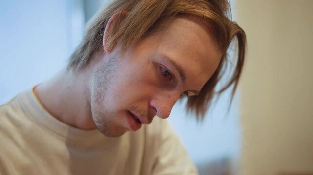 Close up of young man with light brown hair and facial stubble looking downward in soft indoor lighting. subject wears plain white crewneck shirt maintaining consistent positioning