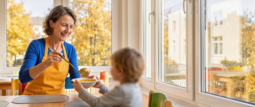 Smiling woman wearing yellow apron using ladle to serve warm pumpkin soup into bowl held by young child. Daycare dining room with large windows showing bright yellow autumn foliage outside