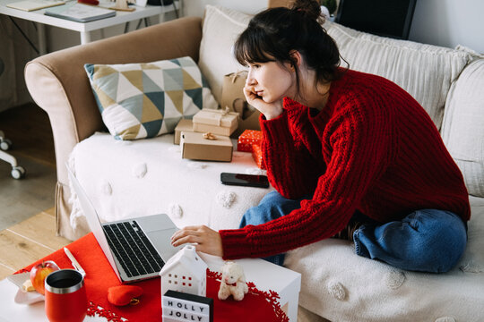 Woman in knitted sweater works on laptop on sofa beside wrapped parcels and HOLLY JOLLY sign. Remote work december, laptop home office christmas, finishing tasks before holidays