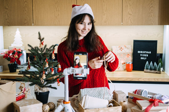 Woman in Santa hat ties raffia ribbon before phone on tripod near CHRISTMAS VIBES ONLY sign. Christmas gift wrapping tutorial, DIY christmas wrapping tips, teaching christmas wrapping technique