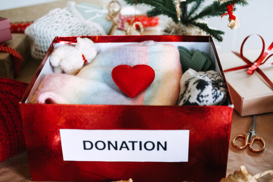 DONATION box packed with knitwear, toy and heart stands among wrapped gifts near Christmas tree. Christmas donation box tree, charity clothing box holiday, giving back christmas season