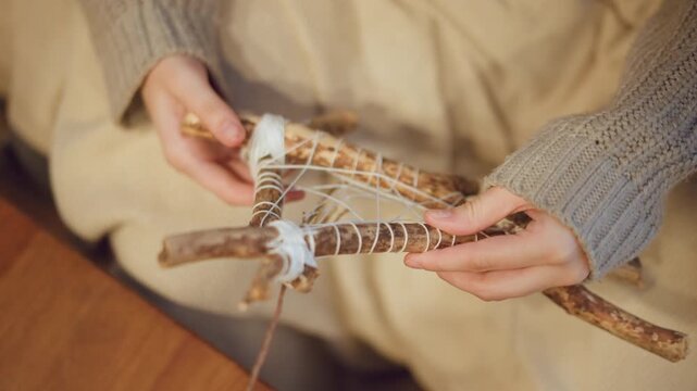 Close up of woman weaving white string around wooden sticks to create intricate web like structure. hands wearing gray knitted sleeves carefully manipulate materials against soft beige