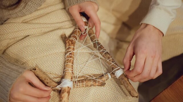 Close up of two people weaving small triangular dreamcatcher with white string around natural sticks on fabric. Hands carefully adjust and tie web like structure showing detailed interaction