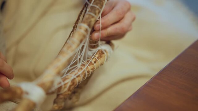 Close up of hands weaving white string around wooden sticks to create small netted structure. process involves carefully tying cord between natural wood pieces forming intricate pattern