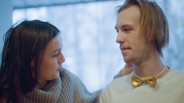 Close up of woman with long dark hair wearing knitted sweater leaning towards man adjusting his golden bow tie. Soft lighting creates warm indoor atmosphere background blurred to emphasize