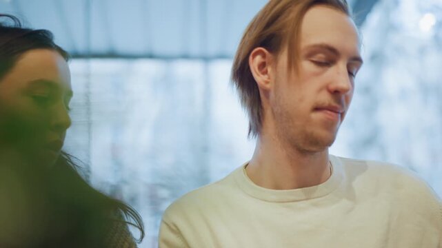 Close up of woman and man sitting side by side in soft lighting both looking away from camera. Woman wears knitted sweater background features blurred greenery and light blue tones