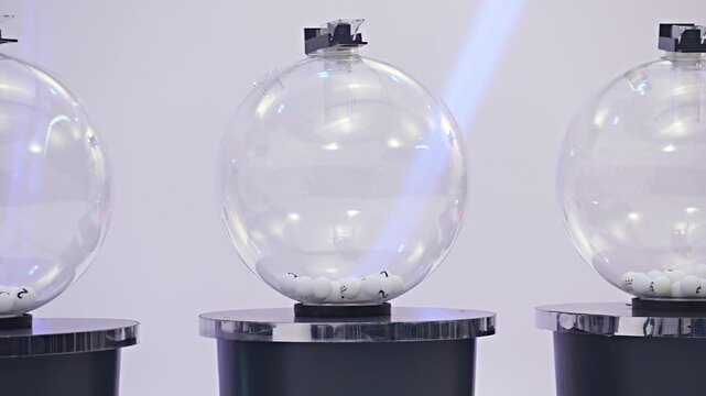 Two transparent lottery raffle drawing spheres with numbered balls on pedestals in bright white studio