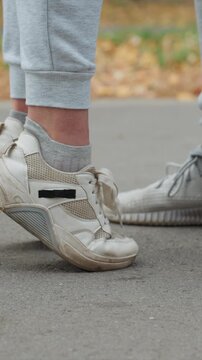 Two pairs of feet in worn sneakers and sweatpants stand. Another identical or similar off white sneaker rests. Autumn foliage appears out of focus behind lower legs shoes