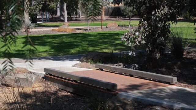 A small footbridge integrated along a paved walking path crossing a dry creek bed inside a leafy suburban park in Melbourne, Australia.