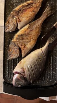 Person using kitchen tongs to flip whole dorado (sea bream) fish while cooking on electric grill pan alongside another fish. Mediterranean seafood cooking, grilling process, healthy dinner preparation