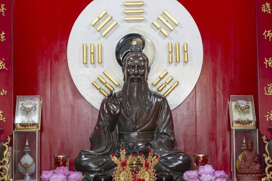 A dark, finely crafted statue of Laozi (Lao Tzu), the legendary philosopher and founder of Daoism, set against a large Yin-Yang symbol and surrounded by the eight Bagua symbols. Red plaques with golden characters and offerings, Chee Chin Khor Temple, Bangkok, Thailand