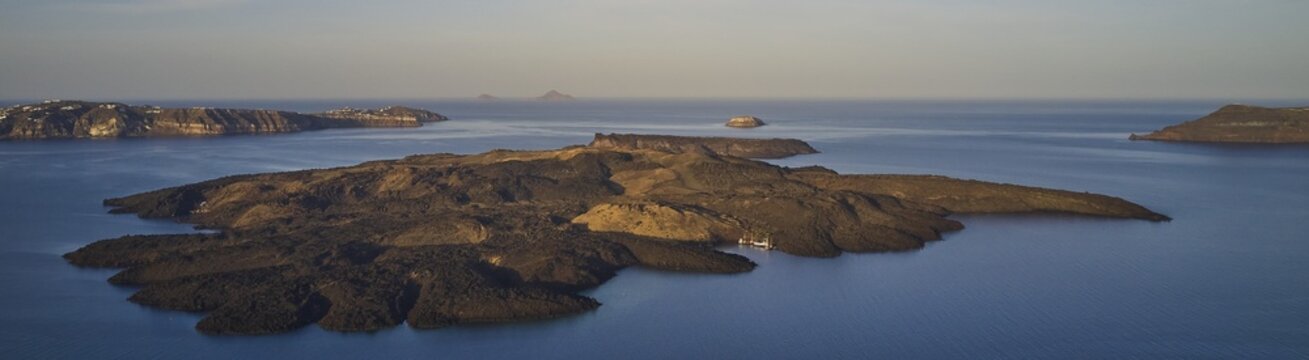 Drone shot, panorama, morning atmosphere, caldera, view of volcanic islands Nea Kameni, Palea Kameni and Aspronisi, view of southern tip of the island, sea smooth as glass and blue, sky light blue and cloudless, caldera in front of Firostefani, Santorini Island, Cyclades, Greece