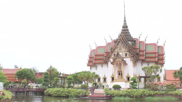 traditional thai architectural structure beside a tranquil canal with a red pedestrian bridge, greenery and landscaped surroundings in thailand