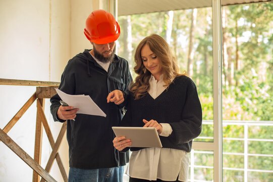 two people, a construction engineer, a man in a helmet and a woman, a customer, an architect, in an unfinished space, checking the quality of the work, calculating, planning, using electronic devices