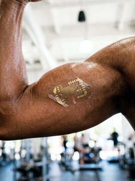 Close up of a muscular male arm with a gold flexible electronic smart patch in a gym, advanced wearable technology, biometric health monitoring and sports science